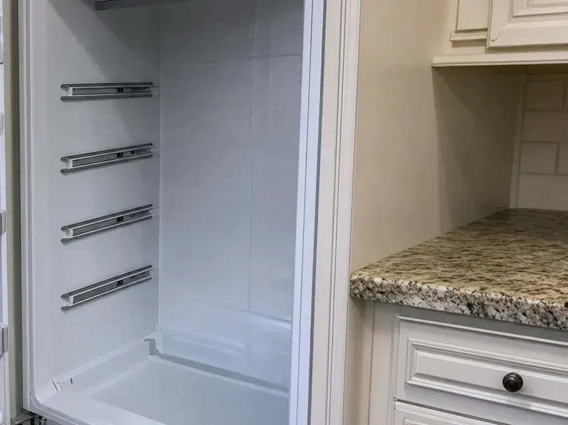 Empty built-in refrigerator compartment with shelf rails, beside cream cabinetry and a granite counter.