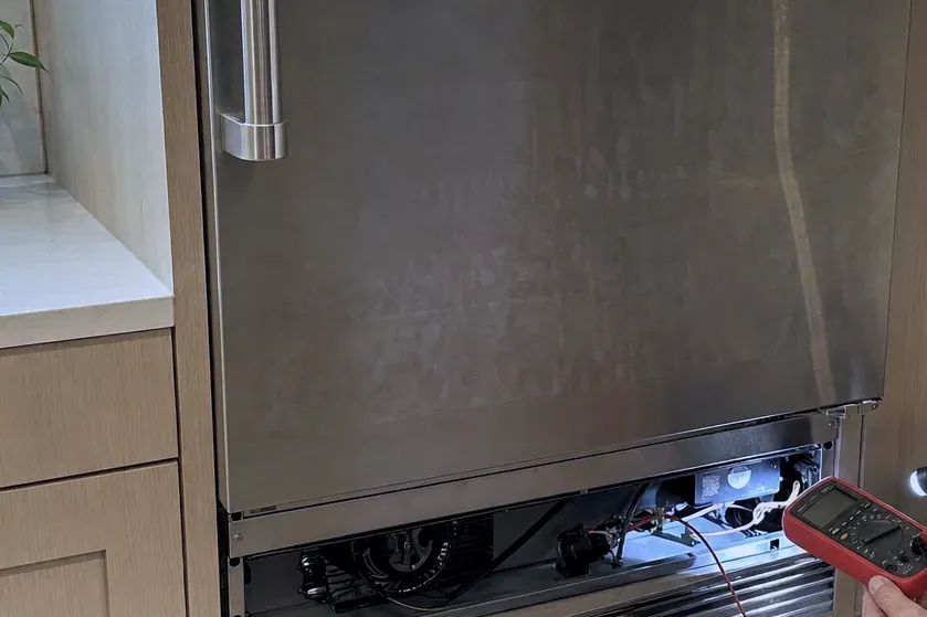 Red multimeter held at the open lower grille of a stainless built-in refrigerator, its fan and wiring visible below the door.